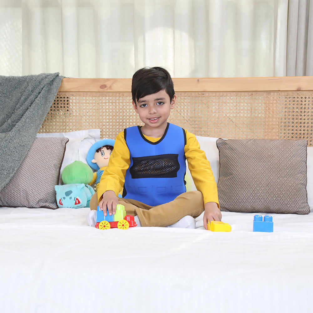 Ableys blue weighted vest for kids worn by smiling boy sitting on bed playing with toy blocks